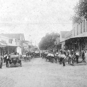 Crowd of white men with cars on town street with buildings on both sides