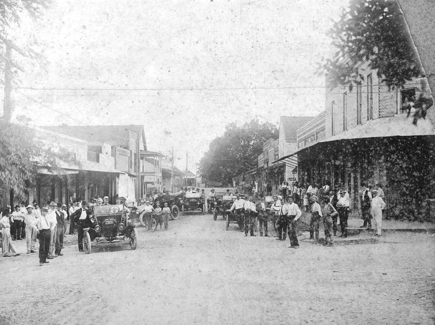 Crowd of white men with cars on town street with buildings on both sides