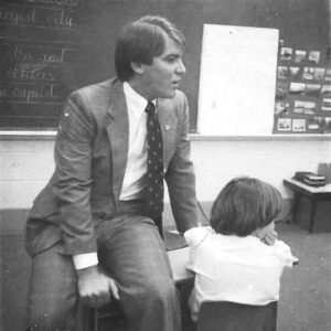 White man in suit and tie sitting on child's desk while speaking in classroom