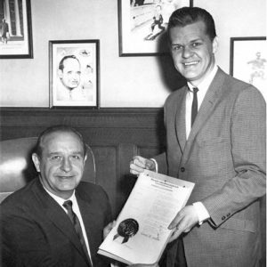 White man in a suit sitting down presenting a document to a younger white man standing up in a suit