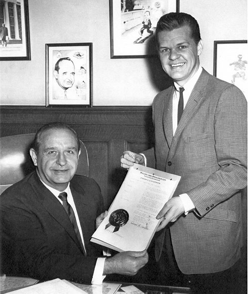 White man in a suit sitting down presenting a document to a younger white man standing up in a suit
