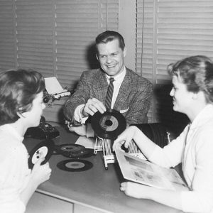 White man in suit and tie showing vinyl records to two young white women