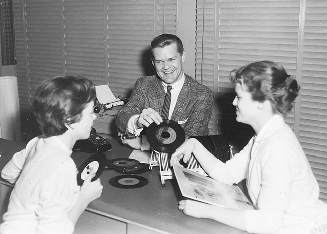 White man in suit and tie showing vinyl records to two young white women