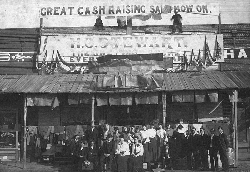 Group of people standing in front of store topped with banner "Great Cash Raising Sale Now On"