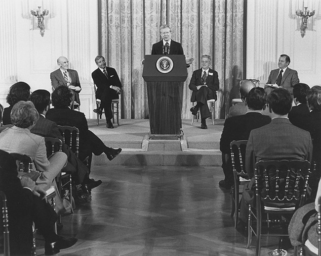 White man speaking at lectern to crowd of white men in suits