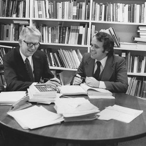 two white men in suits at round table with books in front of bookshelf