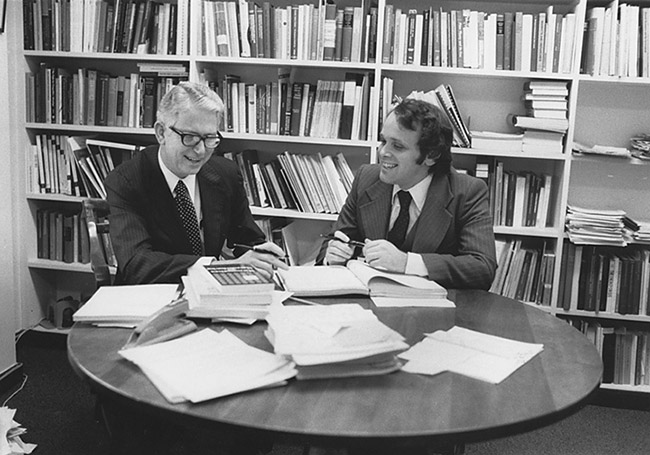 two white men in suits at round table with books in front of bookshelf