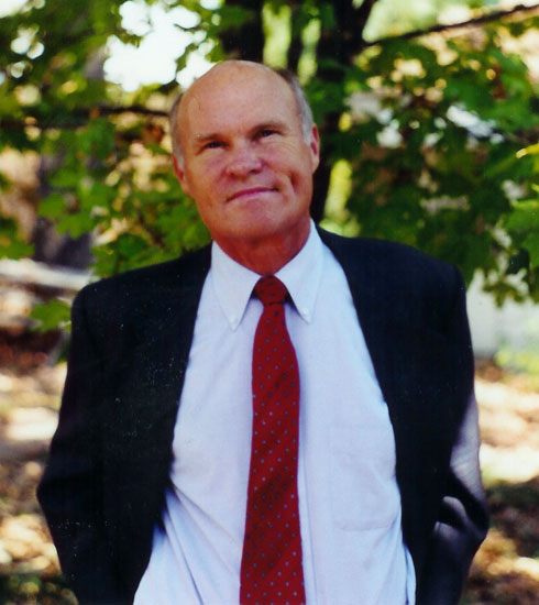 Bald white man in blue suit and red tie with trees in the background