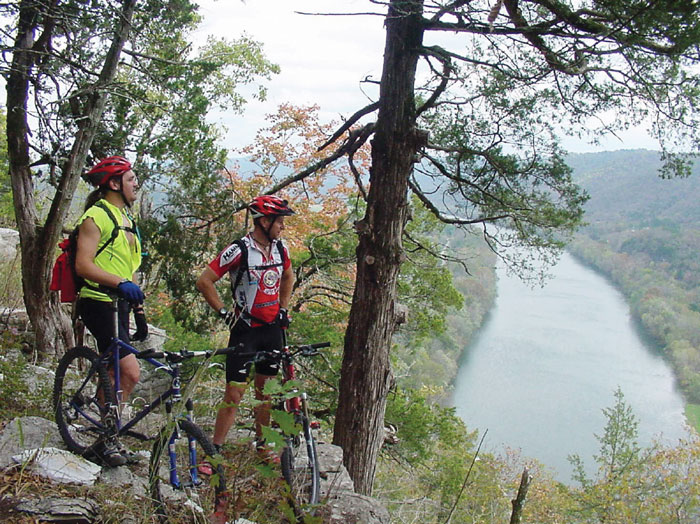 Two white men wearing helmets standing beside bicycles at cliff looking over a river below them