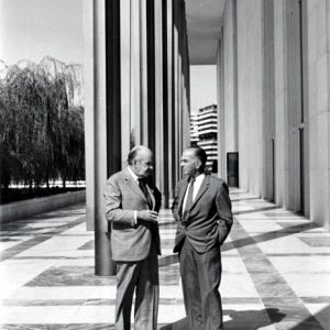 Two white men in suits talking in portico of building with willow trees in background.
