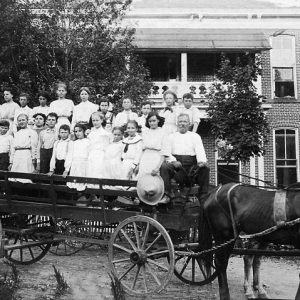 White school children and adults pose on long horse drawn cart outside brick school