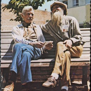 Old white man with mustache sitting on bench next to white man with beard and hat