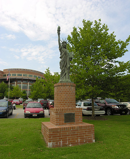 Statue of Liberty replica on brick pedestal on grass with multistory building and parking lot in the background