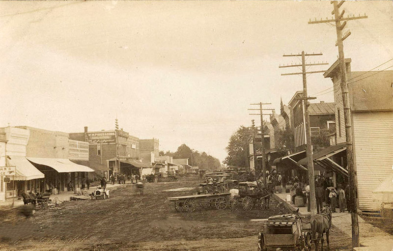 Horse drawn wagons on dirt street outside storefront buildings with power lines