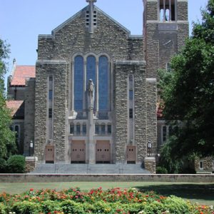 Tall brick church with arched windows and bell tower with trees and flowers in the foreground