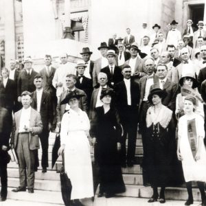 Crowd of white men in suits and women in dresses on the Capitol steps with "Votes for women" pennant flags