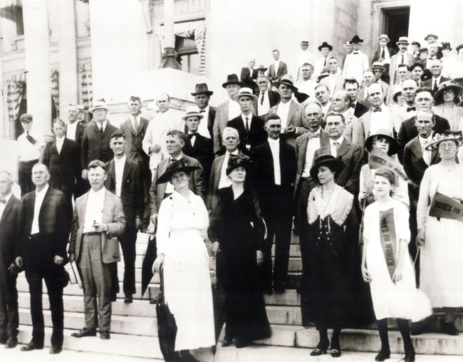 Crowd of white men in suits and women in dresses on the Capitol steps with "Votes for women" pennant flags