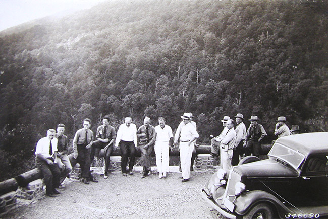 Group of white men and car at scenic overlook with tree covered mountain behind them