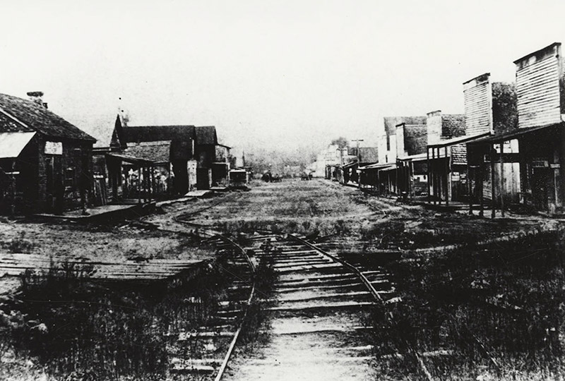 Rows of storefront buildings with covered entrance across from multistory buildings on dirt road with trolley tracks