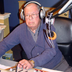 Old white man with glasses and headphones sitting in radio booth