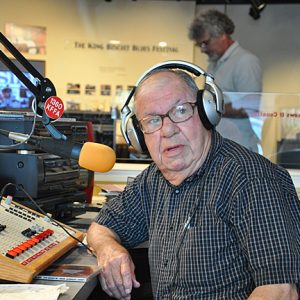 Old white man with glasses and headphones working in radio booth with white man standing in the background