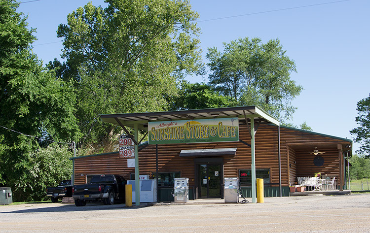 Trucks parked outside building with wood siding and green canopy over two gas pumps with "Sunshine store and cafe" sign