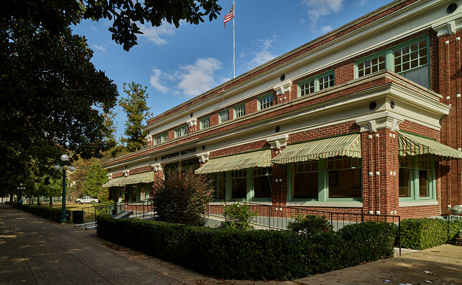 Two-story red brick building with awnings over the first-floor windows and bushes in front