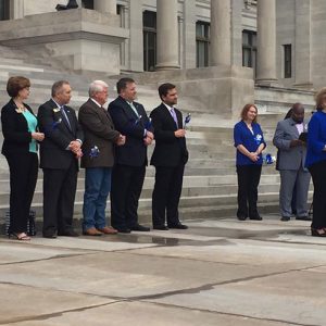 White woman in blue shirt speaking at lectern with mixed crowd behind her on capitol steps