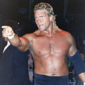 White man with long blonde hair in wrestling shorts pointing off camera