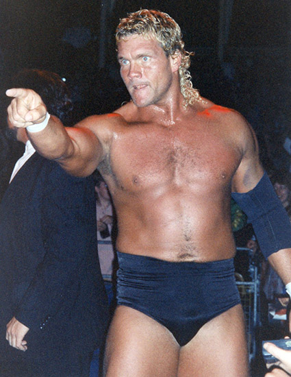 White man with long blonde hair in wrestling shorts pointing off camera