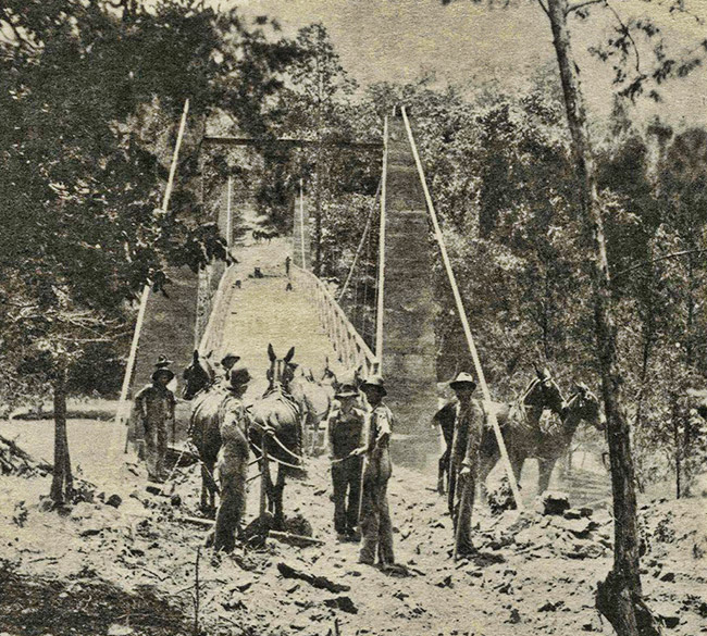 White men and horses at entrance to swinging bridge