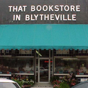 Storefront with large cutout block type sign "that bookstore in Blytheville" awning flowers parked cars