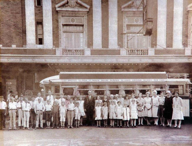 Group of white men women and children posing for a group photo in front of bus outside multistory theater building
