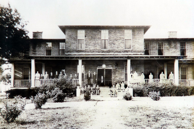 Group of people standing on covered porch of multistory brick house with steps on grass