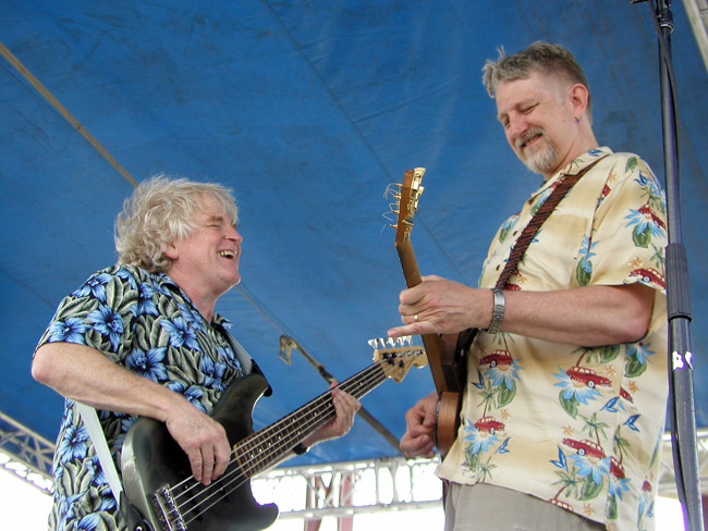 White man singing while playing bass guitar on stage with white man smiling playing electric guitar both under a blue canopy