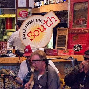 Two white men and African-American man playing instruments inside with "Tales From The South" sign