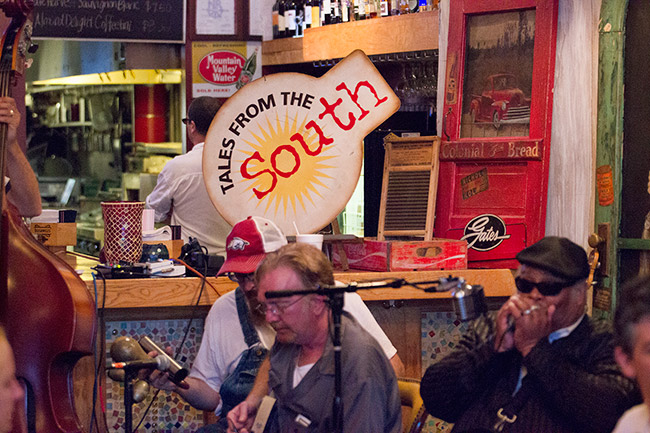 Two white men and African-American man playing instruments inside with "Tales From The South" sign