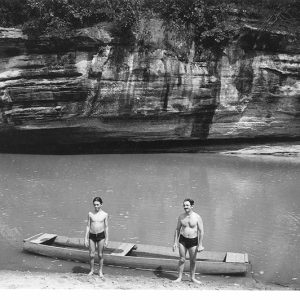 Older white man and son in swimming shorts standing on shore with boat on river