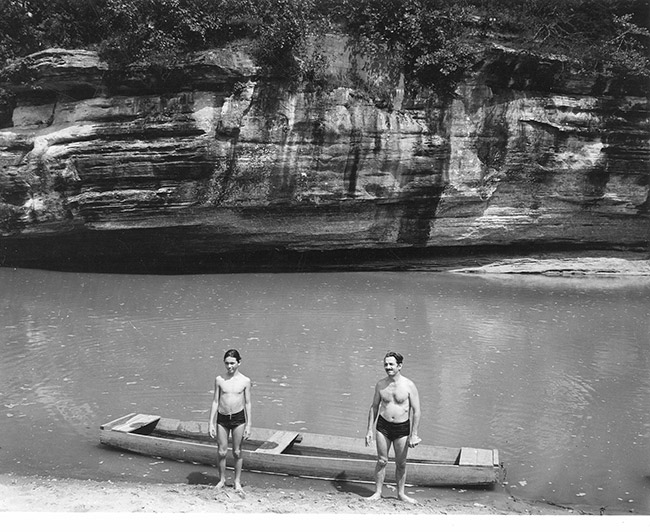 Older white man and son in swimming shorts standing on shore with boat on river