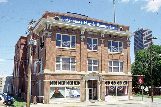 front and side view of three-story brick building with "Arkansas Flag & Banner, Inc." banner and skyscraper in background