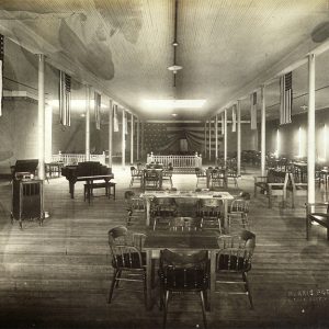High ceilinged interior or room with multiple American flags draped from ceiling above tables, chairs, benches, and a piano