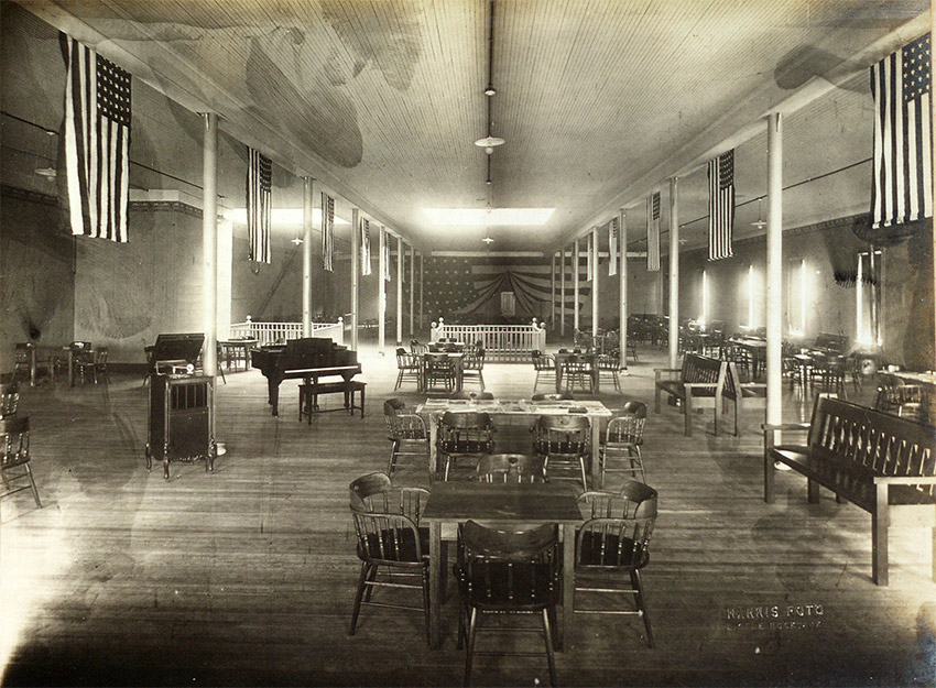 High ceilinged interior or room with multiple American flags draped from ceiling above tables, chairs, benches, and a piano