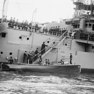 White men in suits and military uniforms boarding naval ship from small boat while sailors stand on its deck