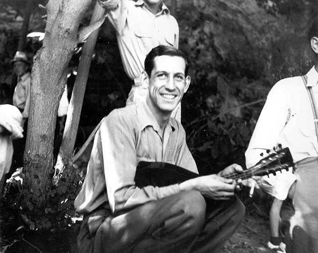 Young white man sitting with guitar amid group of other white men outdoors