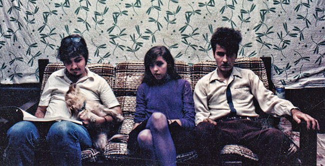 a young dark-haired white woman dressed in purple sits between two young dark-haried white men on a couch with a dog