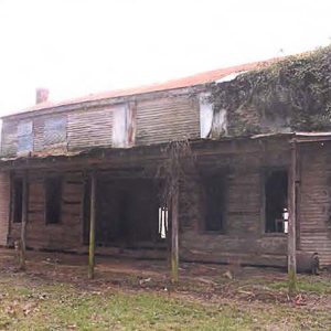 Abandoned and overgrown two-story log house with covered porch on grass