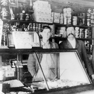 Three white men in suits standing behind counter with display case inside general store with canned items behind them