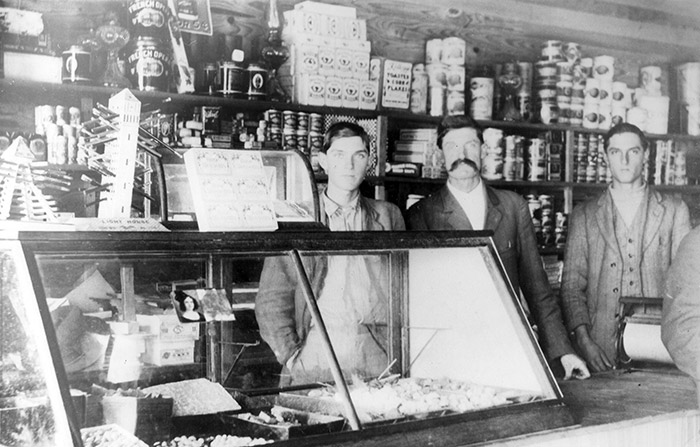 Three white men in suits standing behind counter with display case inside general store with canned items behind them