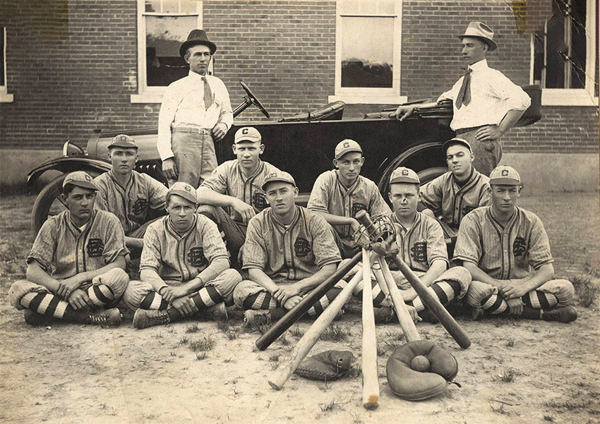 Group of men dressed in baseball uniforms seated on ground with two men in ties standing behind them