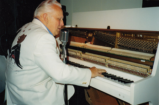 Older white man in white suit jacket with design of a bear in a tuxedo on the back playing piano and singing into microphone